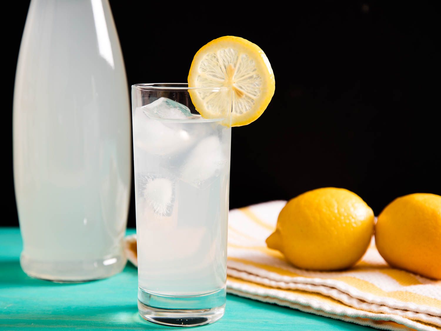 A tall glass of lemonade garnished with a lemon wheel, next to a bottle of lemonade and two whole lemons.