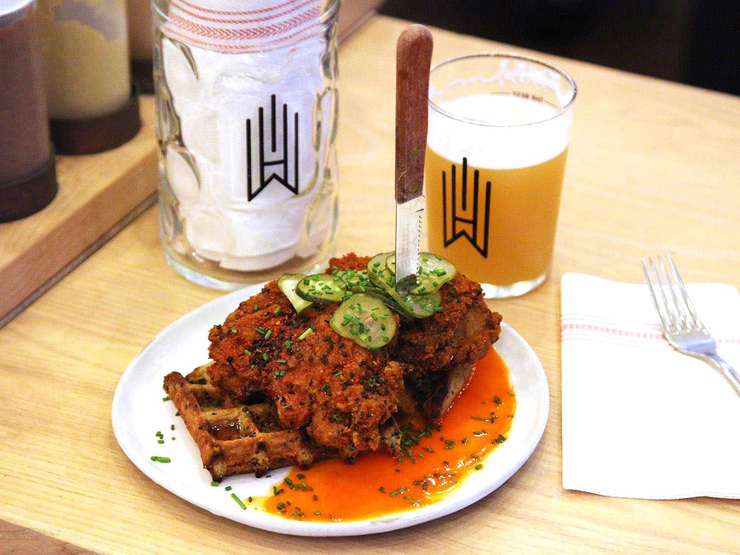 A white plate of kimchi-brined fried chicken atop a cornmeal waffle, topped with dill pickle slices, with a knife stuck vertically into the chicken breast, with a Wursthall glass of beer and another glass containing a napkin in the background.