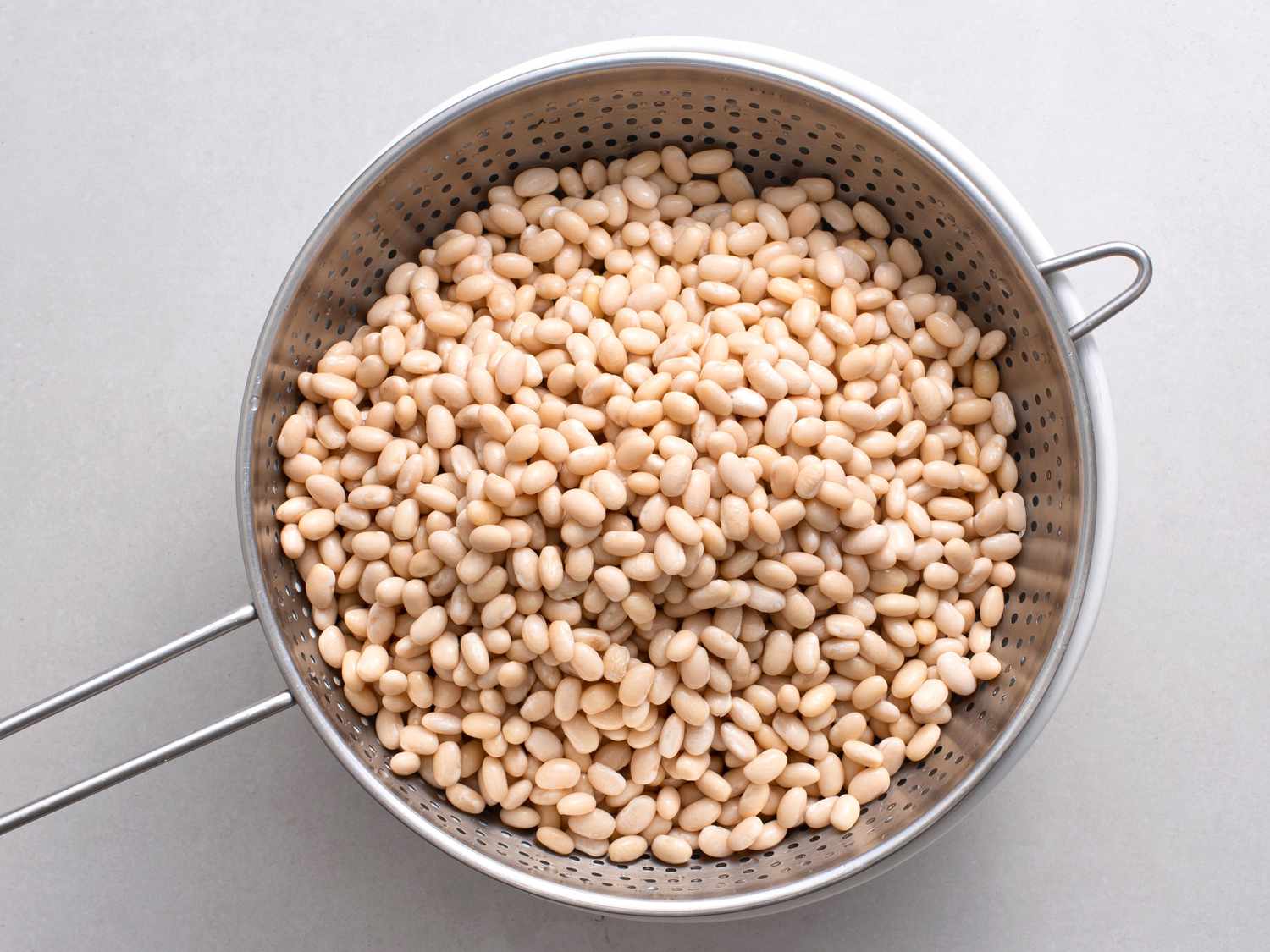 Soaked and drained white beans in a metal colander on a white stone background.
