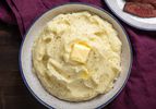 A bowl of fluffy mashed potatoes sits on a table. It's topped with a pat of melting butter and a grinding of fresh black pepper.