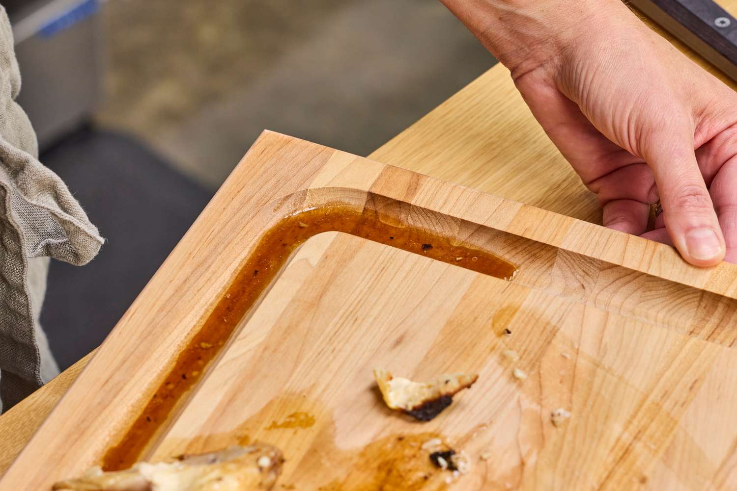 A John Boos Reversible Maple Wood Cutting Board with Juice Groove with a groove containing liquid and food remnants a hand lifting the corner of the board