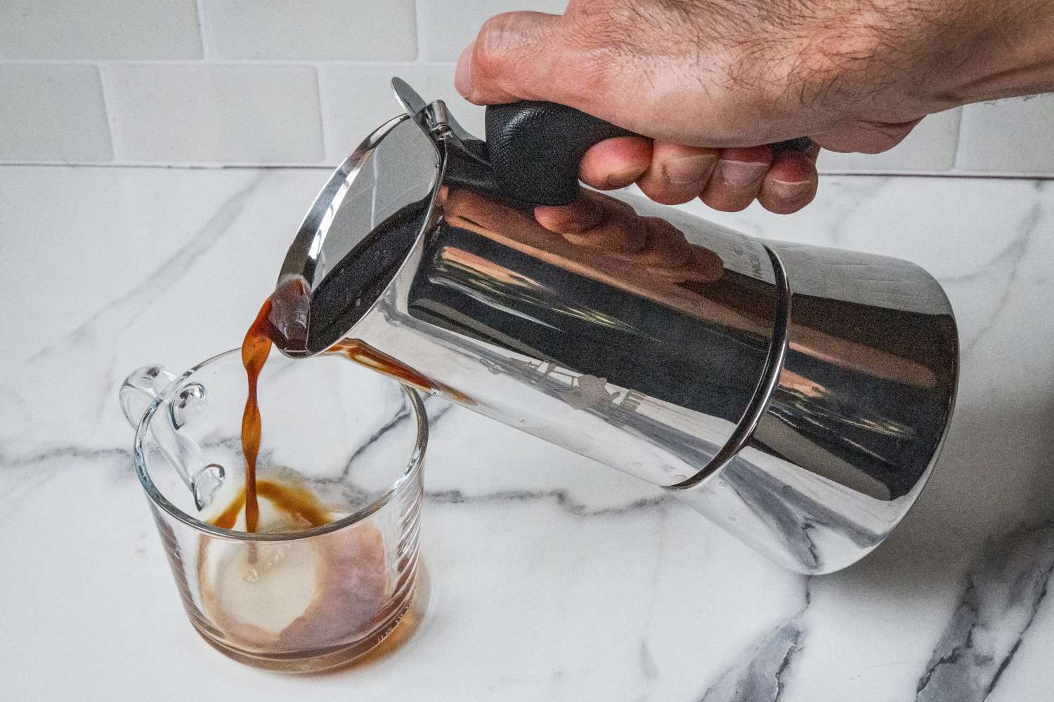 Hand pouring coffee from a stovetop espresso maker into a glass mug