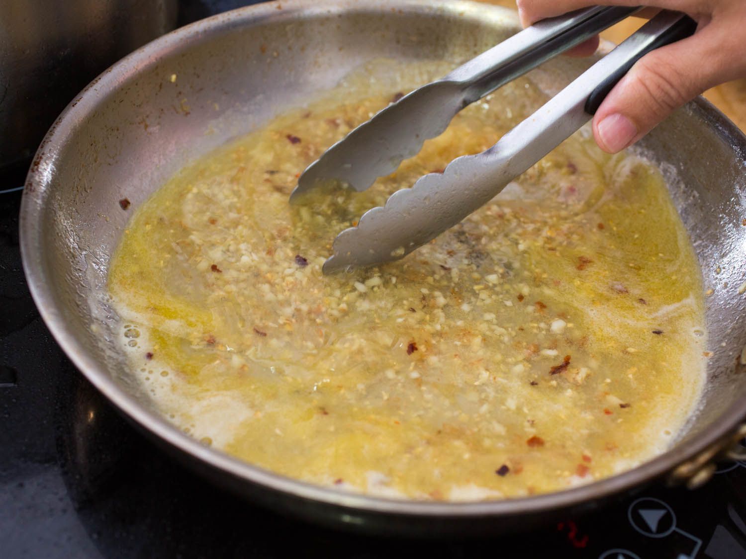 A pair of tongs dipping into a garlic butter sauce in a skillet on the stovetop.