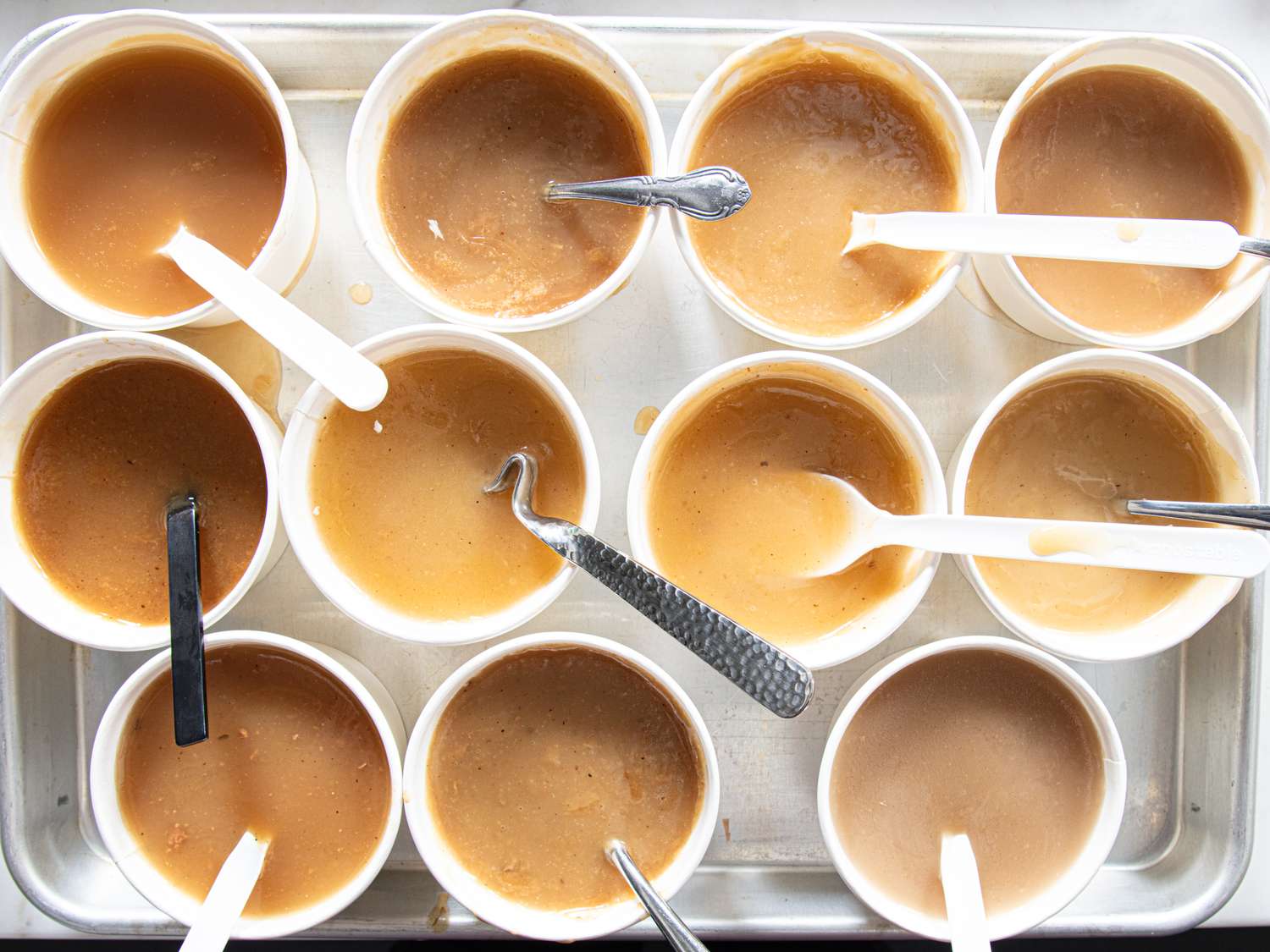 A tray of cups filled with gravy samples each containing a spoon or utensil for tasting