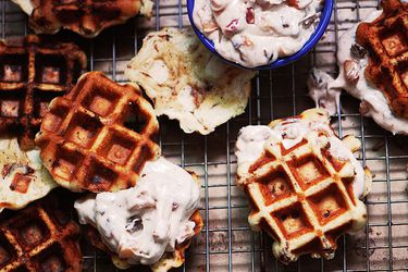 Overhead view of Cinnamon Roll Waffles on a cooling rack over a rimmed baking sheet. A ramekin of Bacon-Apple Frosting is in the frame and one of the waffles has been frosted.