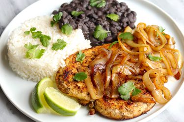 A plate of Cuban-style pollo a la plancha topped with caramelied onions, and served with white rice, black beans, lime wedges, and cilantro.