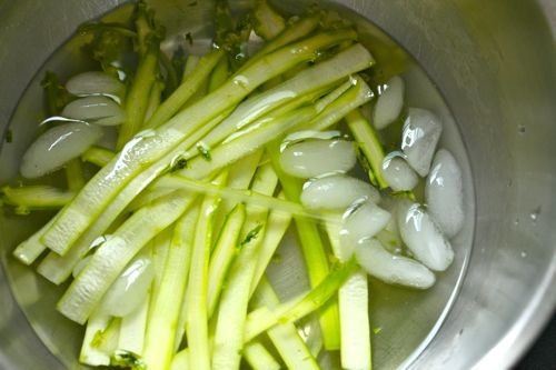 Thin slices of raw asparagus resting in a metal bowl filled with ice and water.