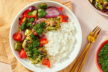 A dish with grilled chicken thighs vegetables chimichurri sauce and white rice on a plate utensils and a sauce bowl beside it