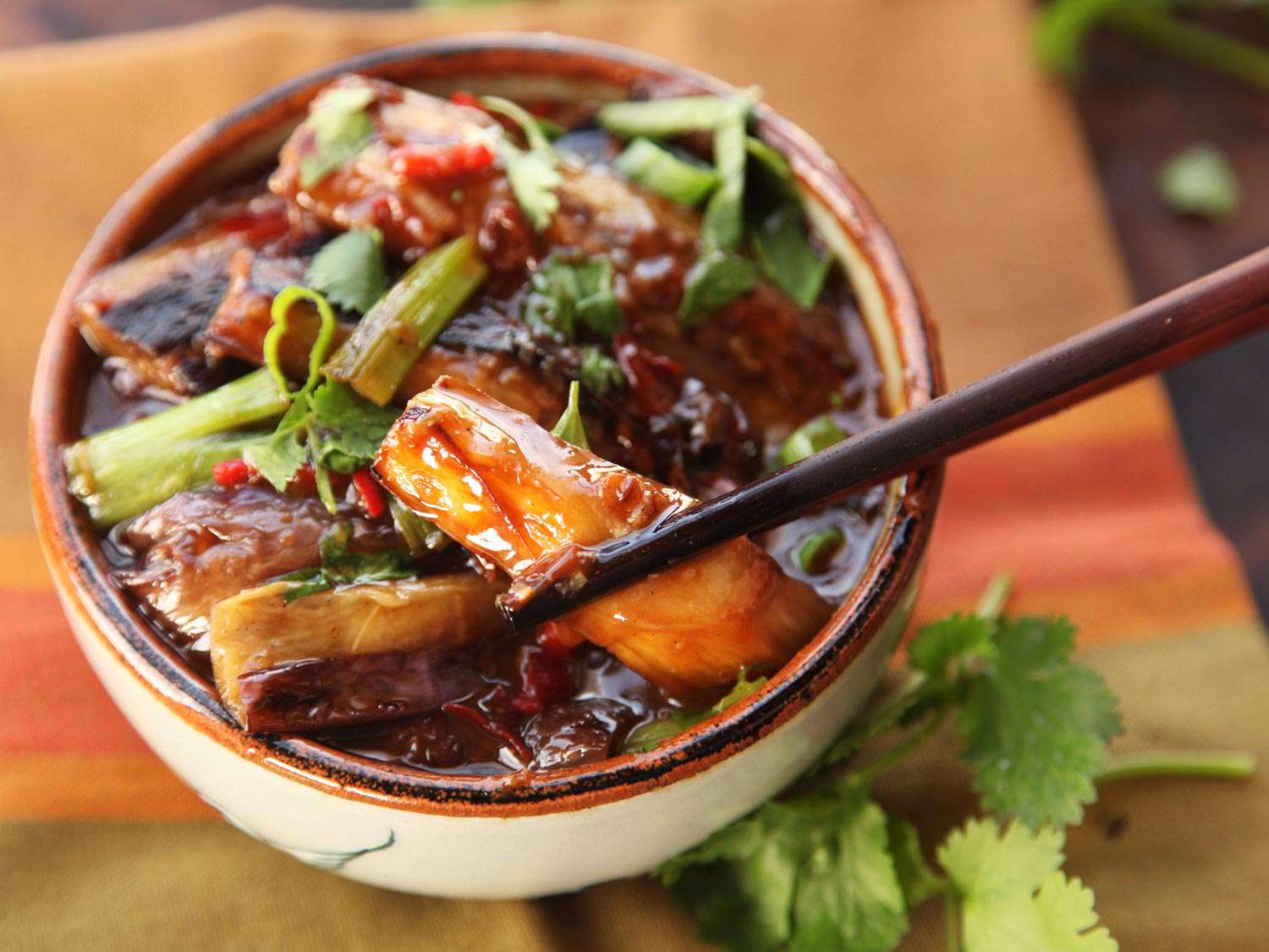 A piece of eggplant is raised from the serving bowl with a pair of chopsticks.
