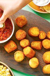 An oval wooden platter holding crispy deep-fried jalapeÃ±o poppers and a small bowl of tomato sauce. There is a hand dipping a popper into the tomato sauce, and the periphery of the image contains other snacks, such as pretzels, popcorn, and chips.