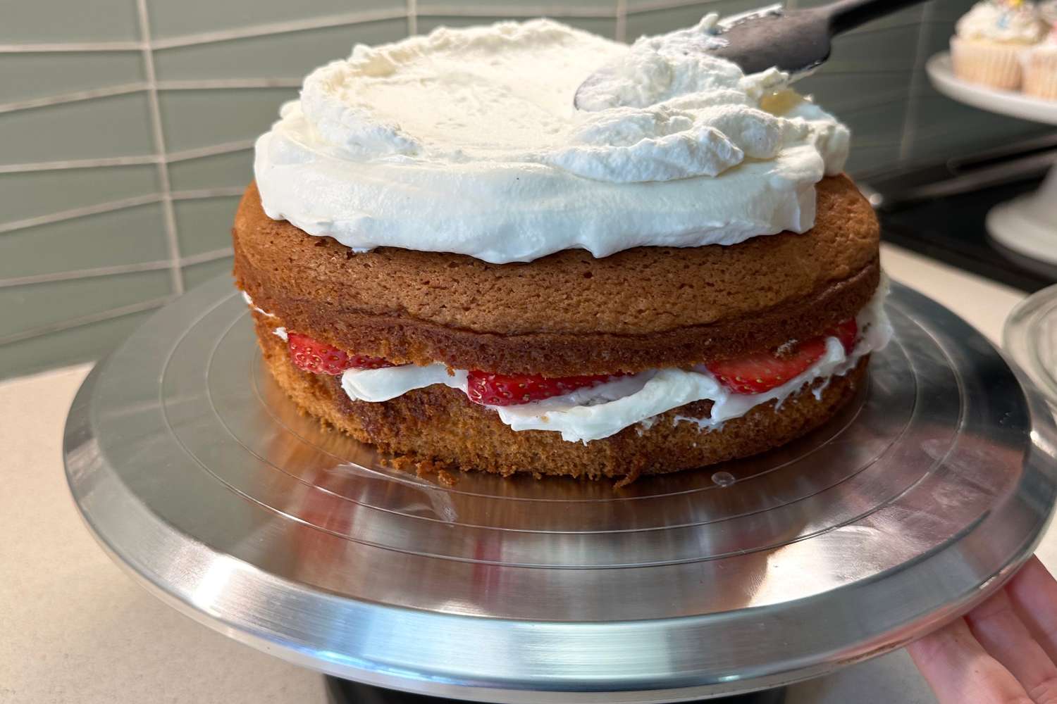 A strawberry cake is frosted with whipped cream on a revolving cake stand