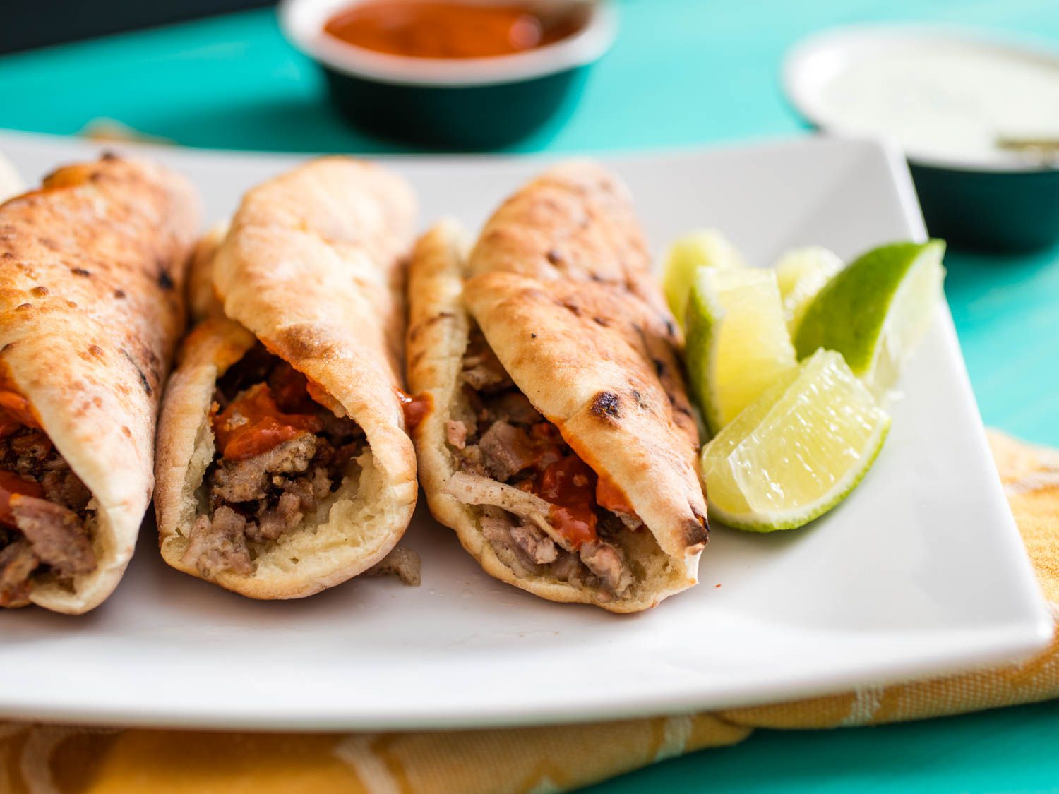 Close-up of a platter of tacos árabes, flanked by lime wedges. Chipotle salsa and yogurt sauce lurk in the background.