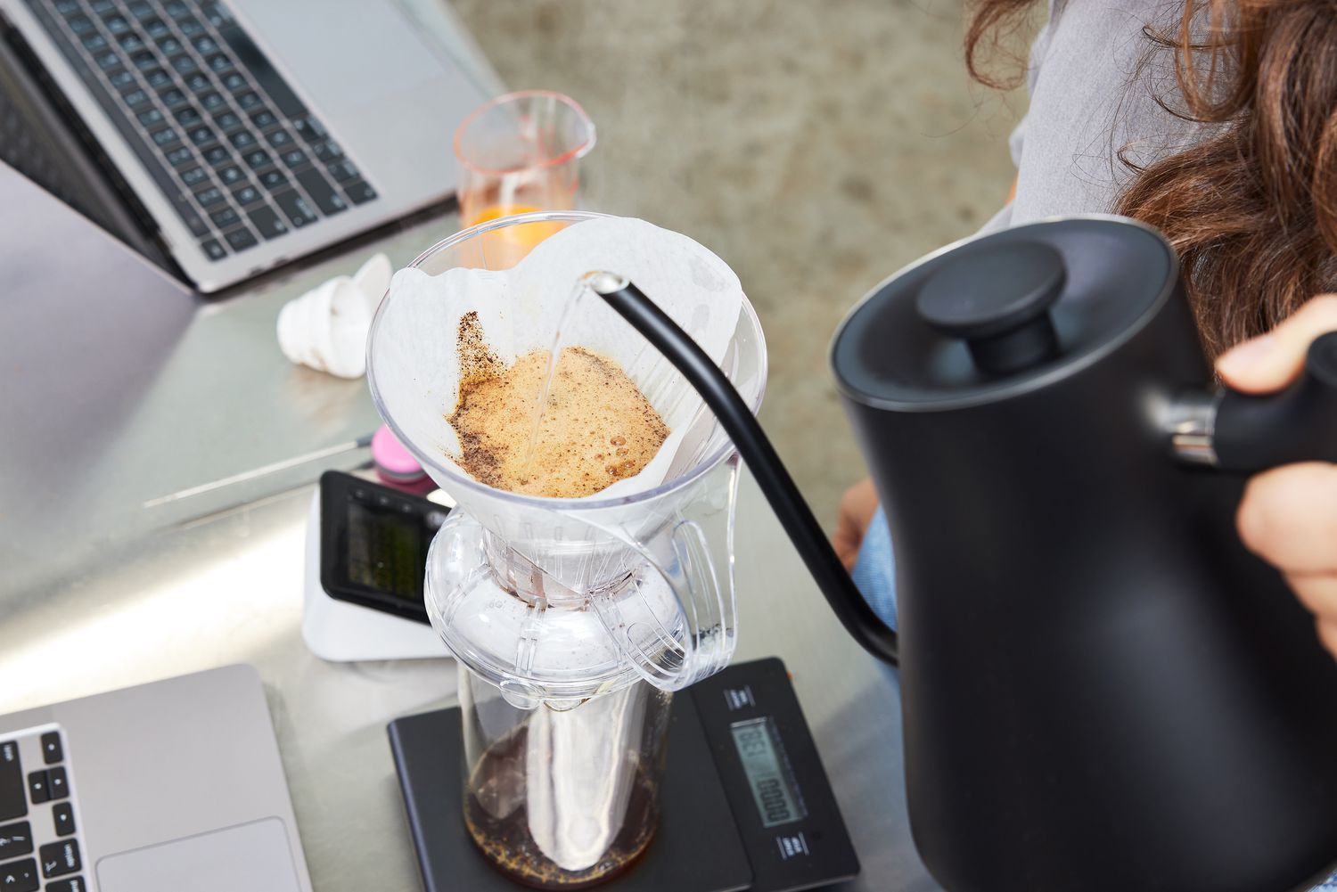 A person pouring water into a Clever Dripper coffee maker on a scale, tools including a laptop and thermometer on the table