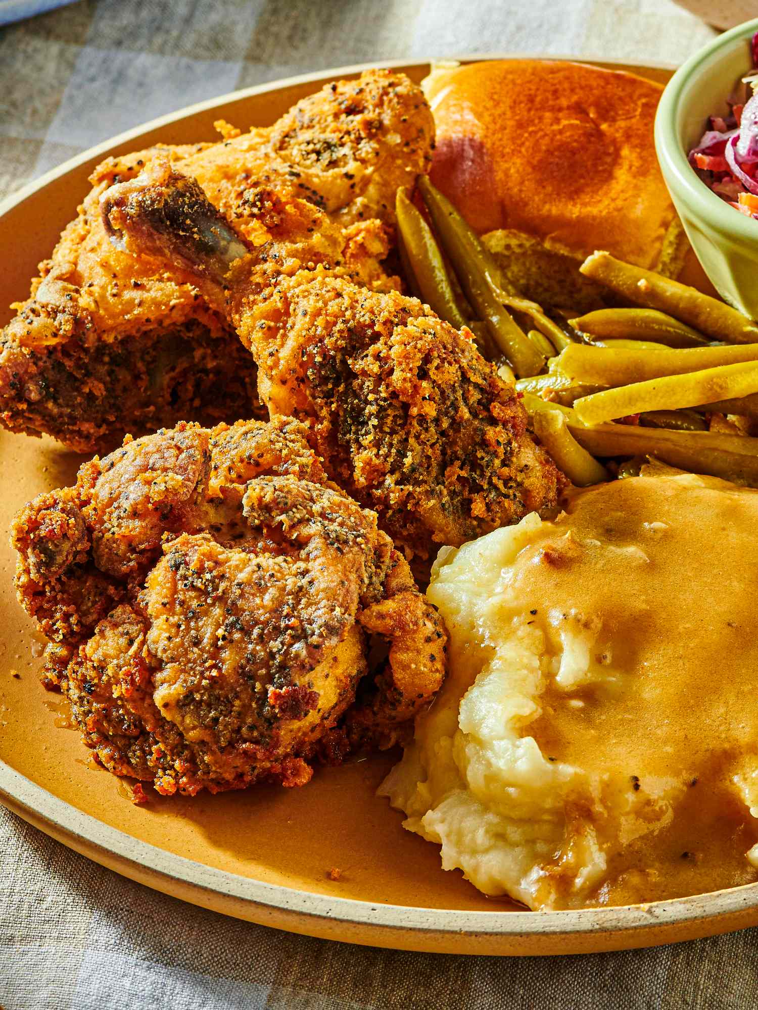 Dish of fried chicken with coleslaw, greenbeans, and mashed potatoes, with a fork and serving dishes in the background