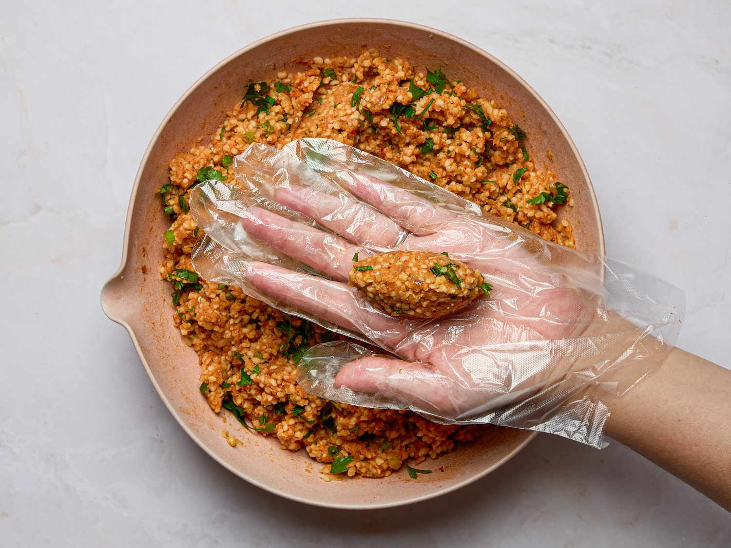 A hand wearing a glove shaping lentilbased mixture into a small oblong piece above a bowl of similar mixture