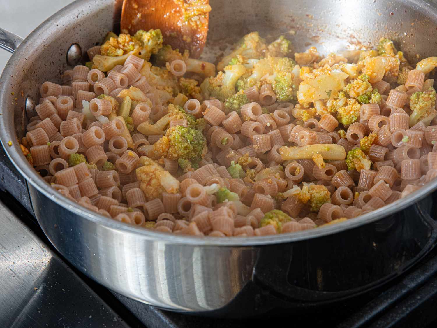 A skillet containing pasta mixed with pieces of romanesco and sauce being stirred with a wooden utensil