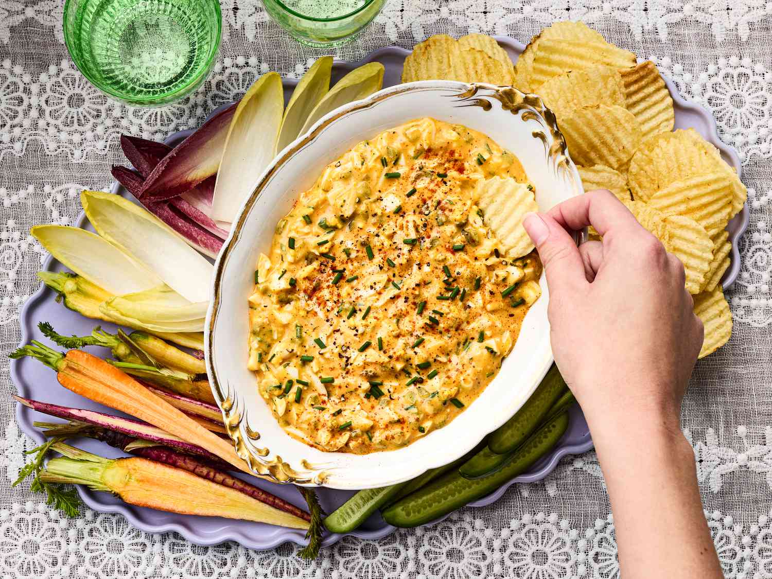 A hand dipping a potato chip into deviled egg dip, surrounded by vegetables on a tray