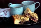 Two slices of blackberry thyme crumb cake, served on a black plate with two mugs of coffee.