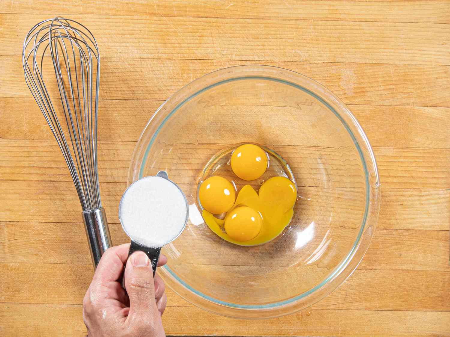 4 eggs in a glass bowl with a hand about to add sugar, a whisk nearby.