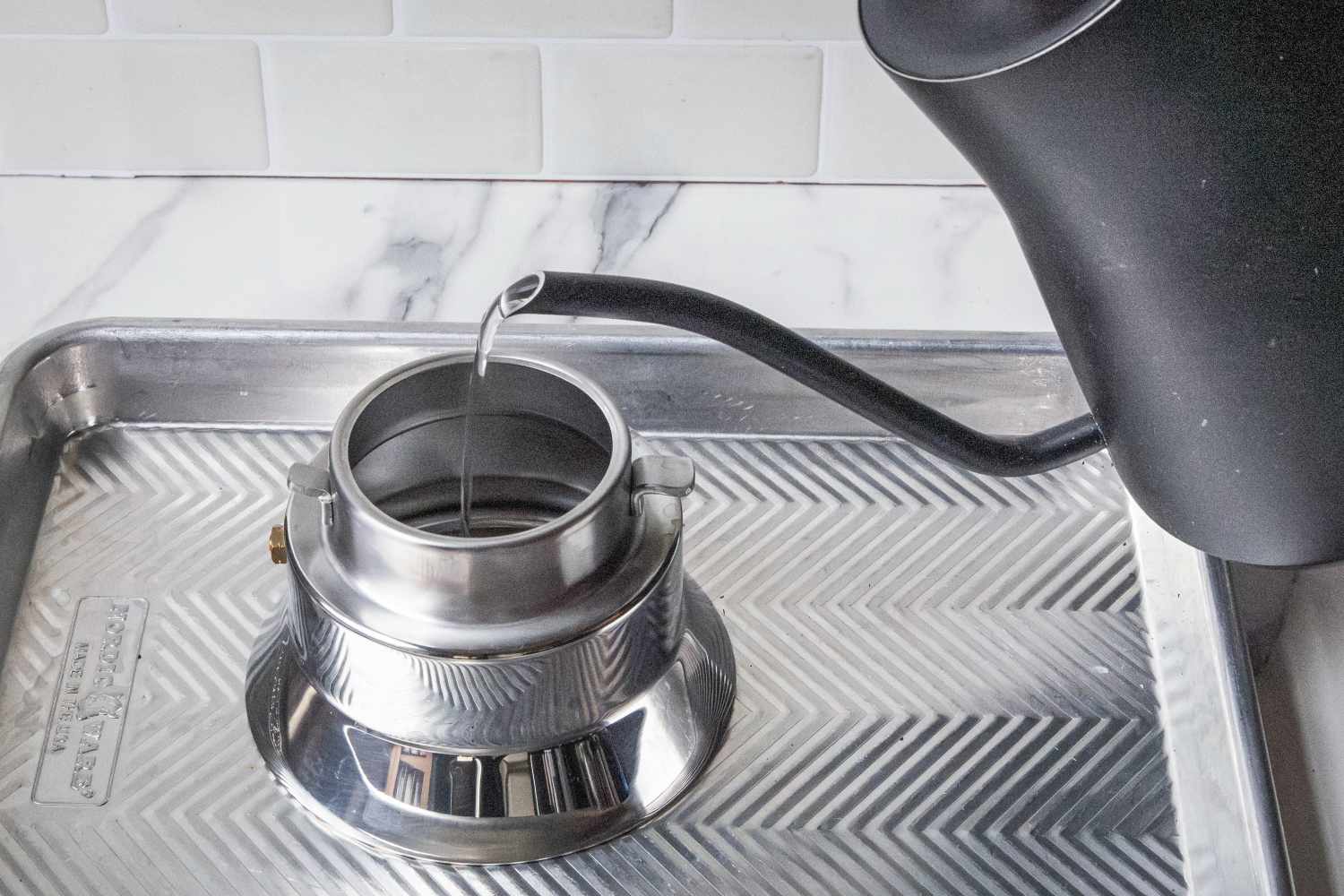 Pouring water into a metallic container using a gooseneck kettle placed on a patterned tray