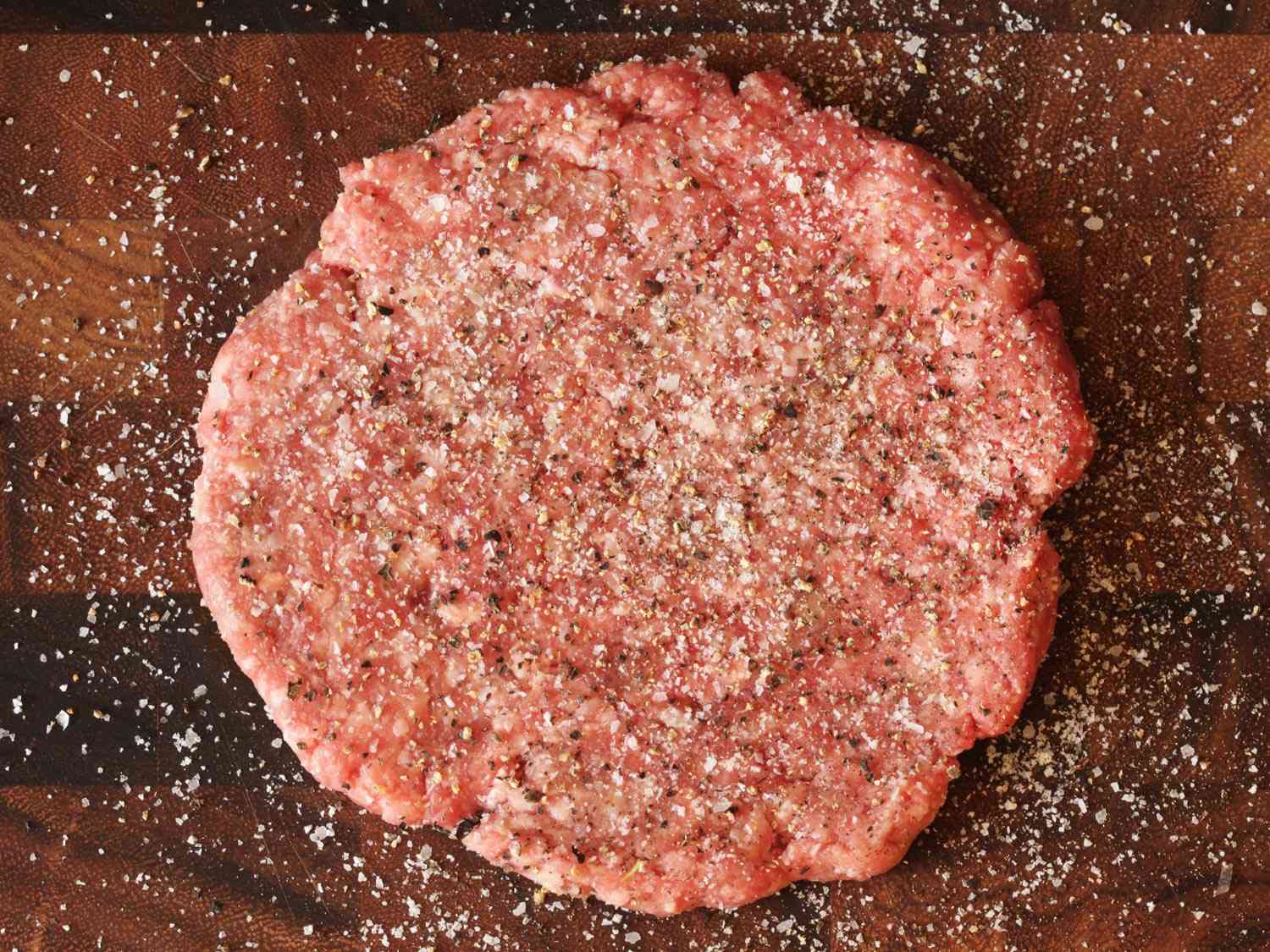 A raw hamburger patty, liberally seasoned with salt and black pepper.