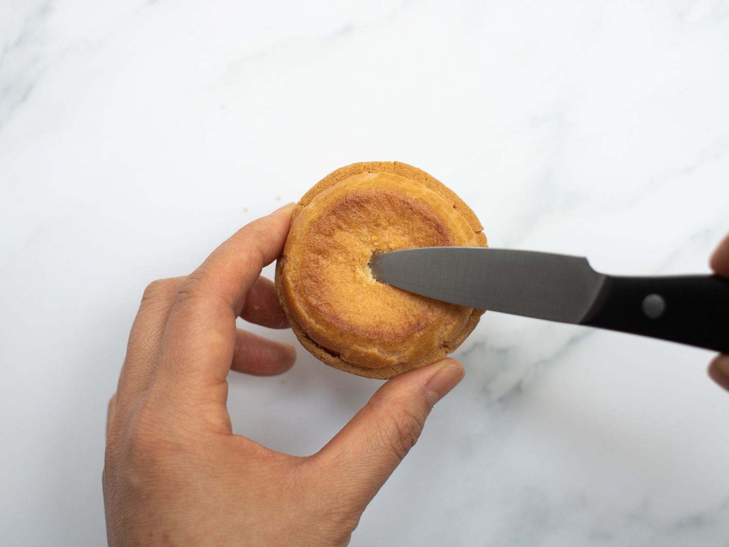 A knife cutting a hole into the bottom of a choux au craquelin for access to place filling in.