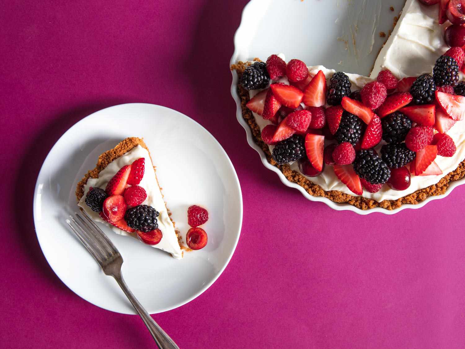 Overhead shot of the berry-studded cheesecake and a plated slice on a magenta table cloth.