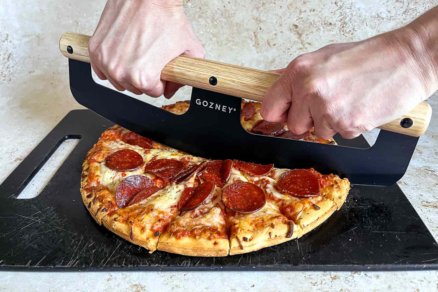 Hands slicing a pepperoni pizza using a Gozney pizza rocker on a cutting board