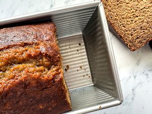 Loaf of banana bread in a metal baking pan on a marble countertop with slices of bread visible nearby