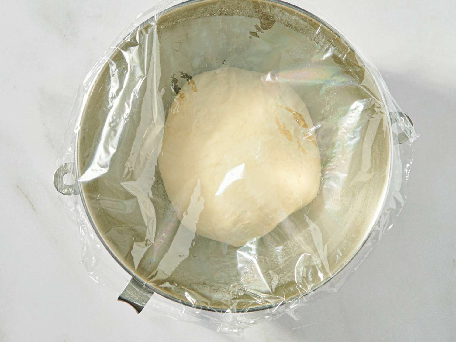 A dough ball covered with plastic wrap in a mixing bowl set for rising