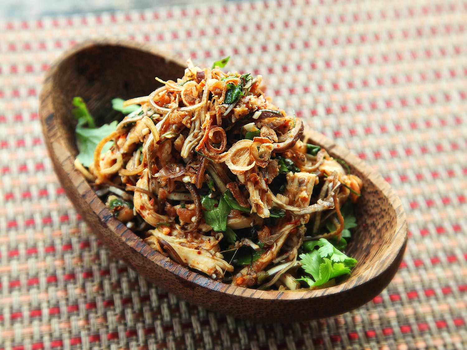 A wooden boat-shaped dish holding a Thai salad of poached chicken, banana blossom, fried shallots, fried garlic, fried lemongrass, and spicy dressing