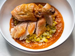 Closeup of a bowl containing sliced roast chicken pieces served atop a puddle of romesco crowned with a smattering of creamy corn.