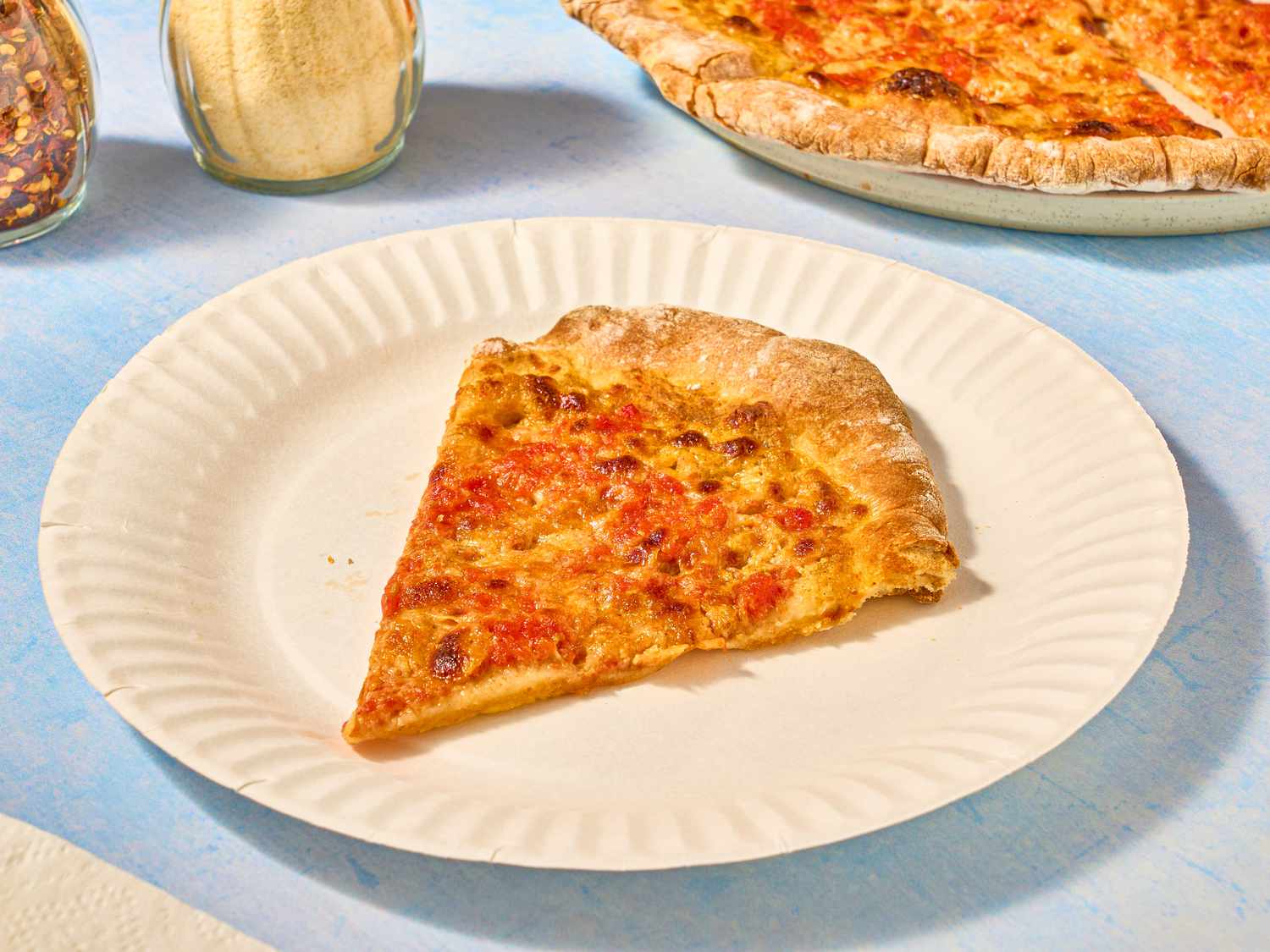 slice of mustard pizza on paper plate, and whole pizza in back, with parmesan and red pepper shakers 