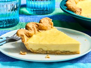 Slice of custard pie on a white plate with a fork next to it, surrounded by blue glasses and a colorful tablecloth