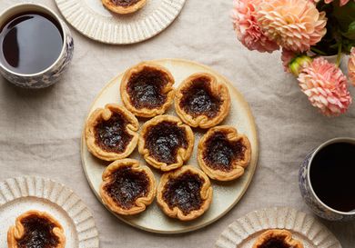 Overhead of platter with Canadian butter tarts. 3 smaller plates have one tart each. Coffee and flowers on the tabletop, with linen tablecloth.