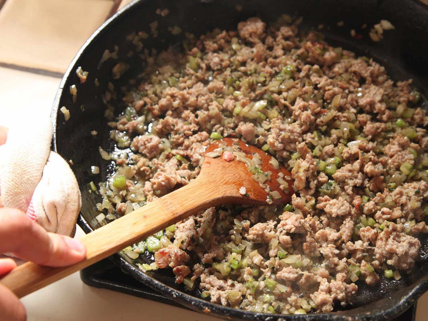 Using a wooden spoon to stir sausage, celery, onions, sage, and garlic in a cast iron skillet