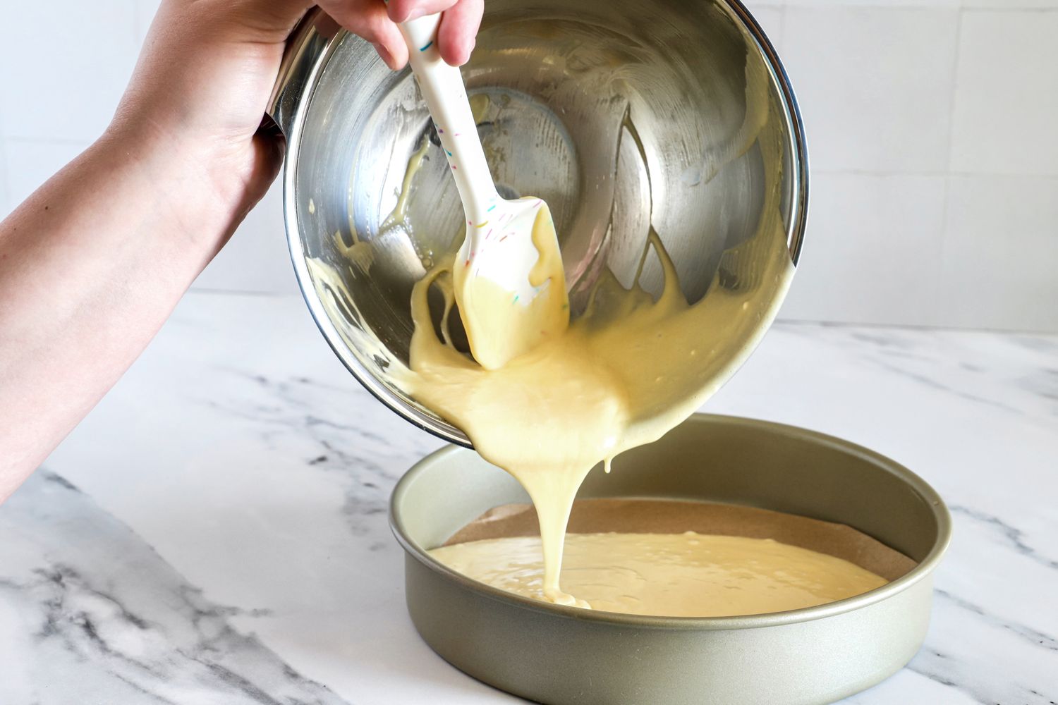 Cake batter being poured from an All-Clad mixing bowl into a baking pan by hand with a spatula