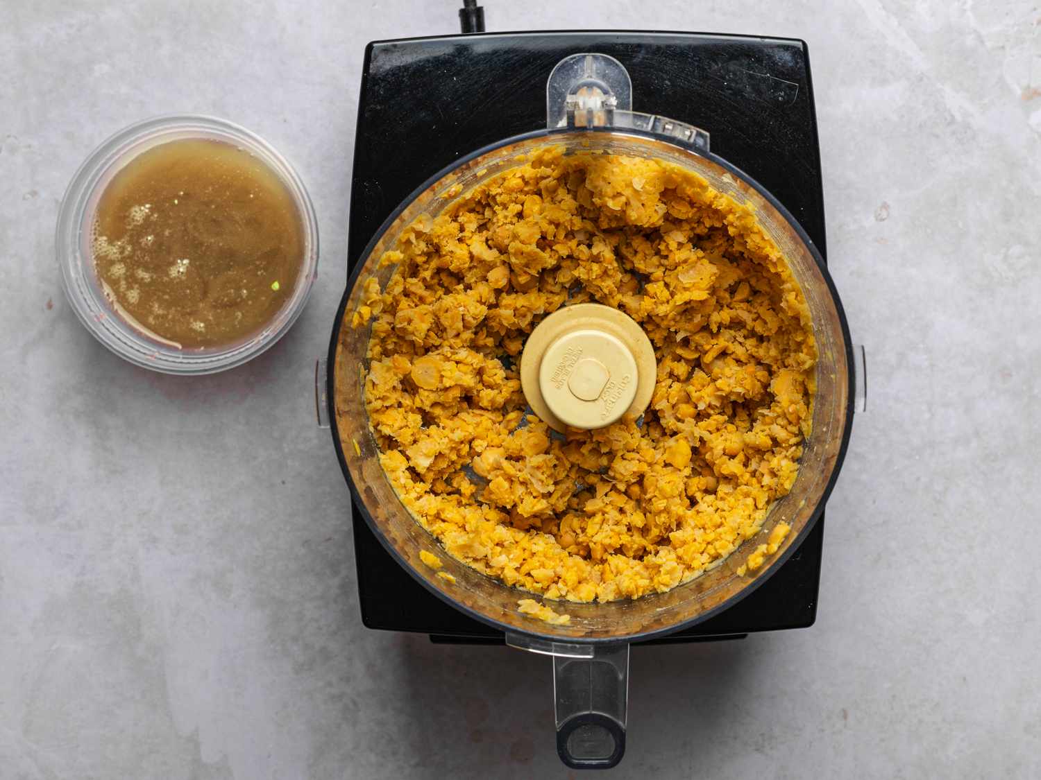 Drained peas, with the liquid in a small cup to the left, pulsed until roughly chopped inside a food processor bowl.