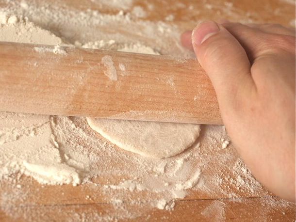 A wonton wrapper being rolled on a heavily floured cutting board by a rolling pin held by a hand.