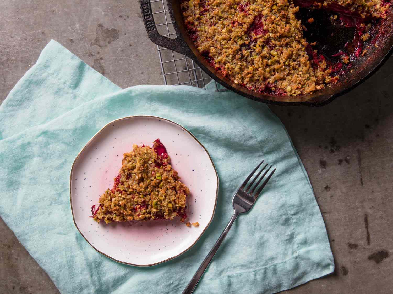 Overhead view of creamy beet gratin with pistachio crumble, served on a small plate. A skillet full of the gratin is nearby.