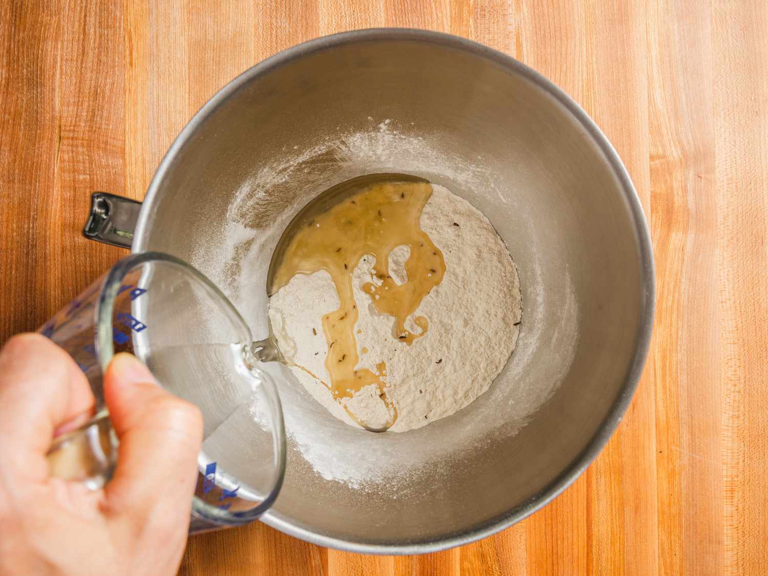 Overhead view of dry mix for samosa dough in a stand mixer bowl with oil being poured from a measuring cup into the bowl.