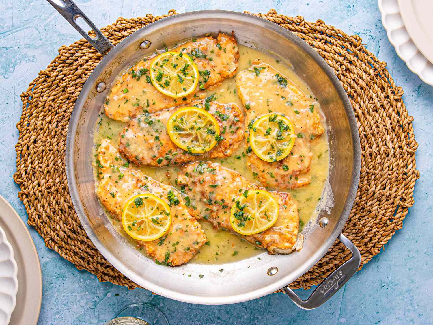 A dish of chicken Francese in a pan with lemon slices and sauce on a woven placemat on a light blue surface