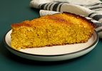A slice of Southern-style unsweetened cornbread on a round white ceramic plate with a dark green rim. The cornbread is resting on a dark teal surface and there's a dishcloth in the background.