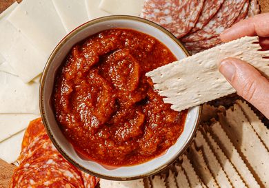 A hand holding a cracker above a bowl of red dip surrounded by slices of cheese and meats