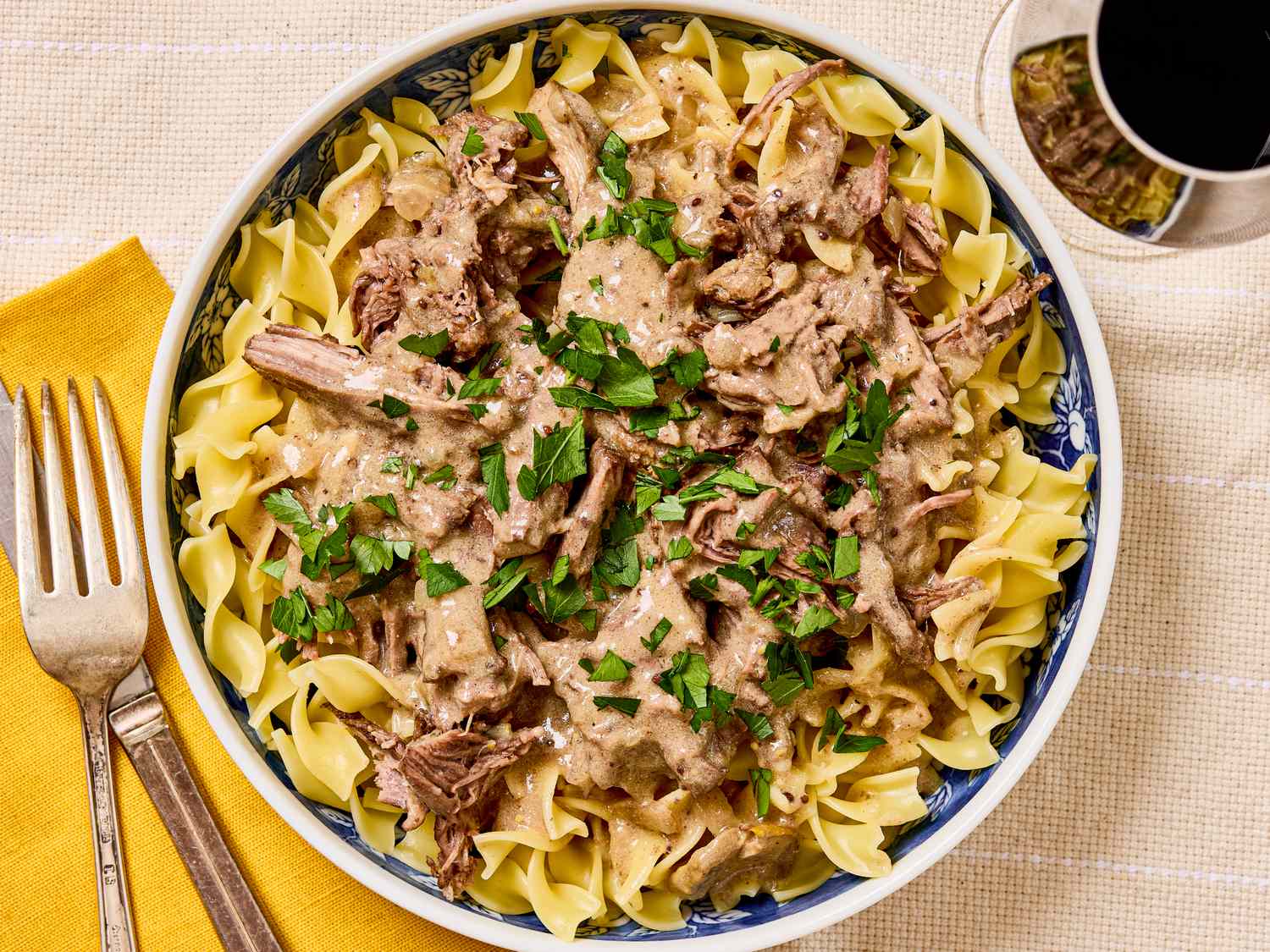 A dish of beef stroganoff served over noodles garnished with parsley and displayed in a bowl