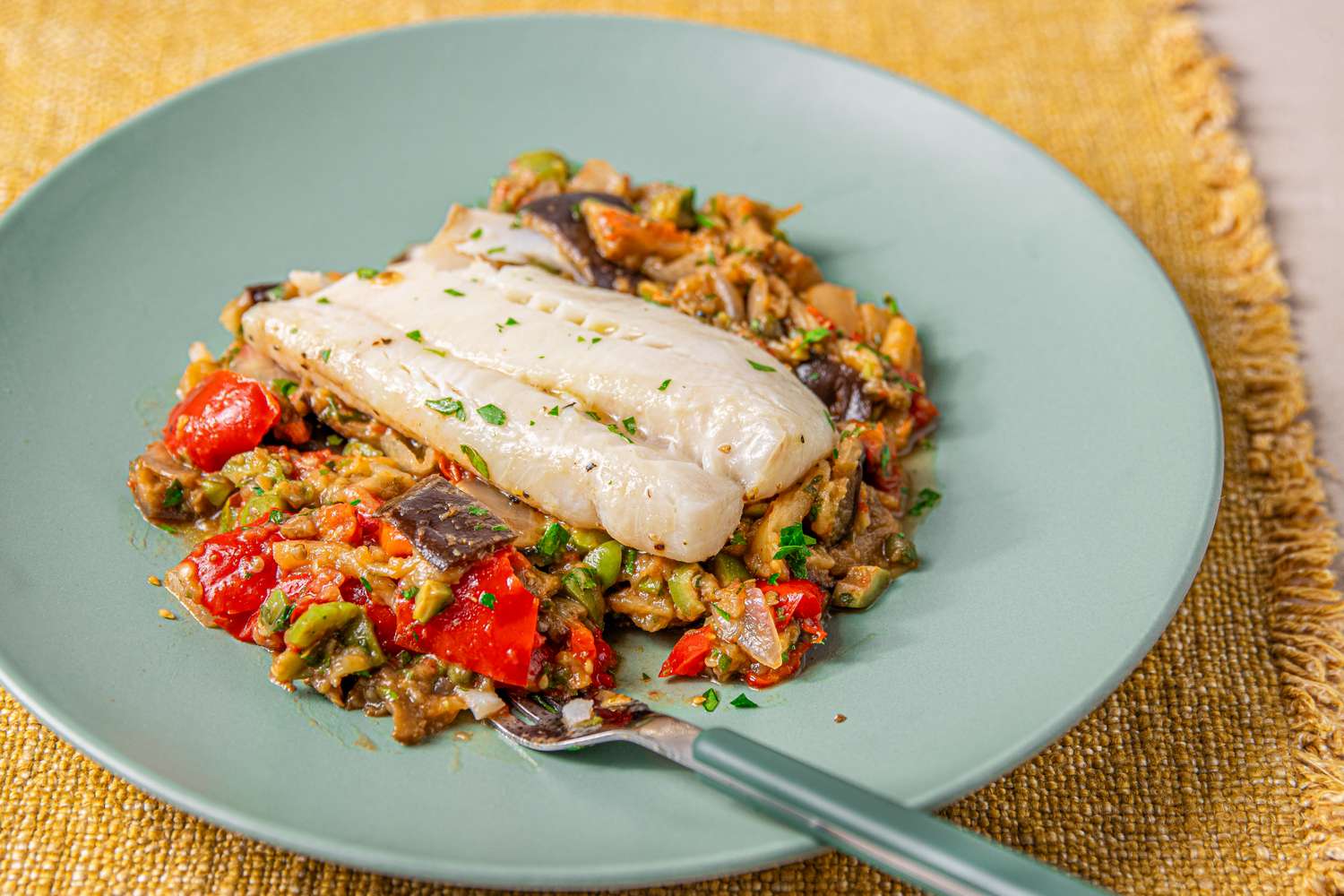 A plate of cod fillet served on a bed of caponata placed on a greencolored plate with a fork