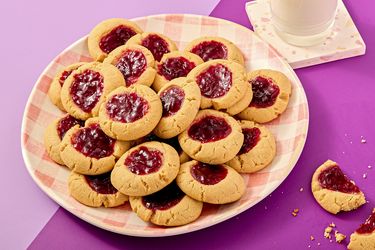 A plate of thumbprint cookies with jam filling next to a glass of milk and one partially eaten cookie