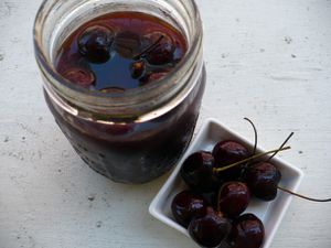 An opened jar of homemade brandied cherries in syrup accompanied with a small bowl of stemmed fresh cherries on the side.