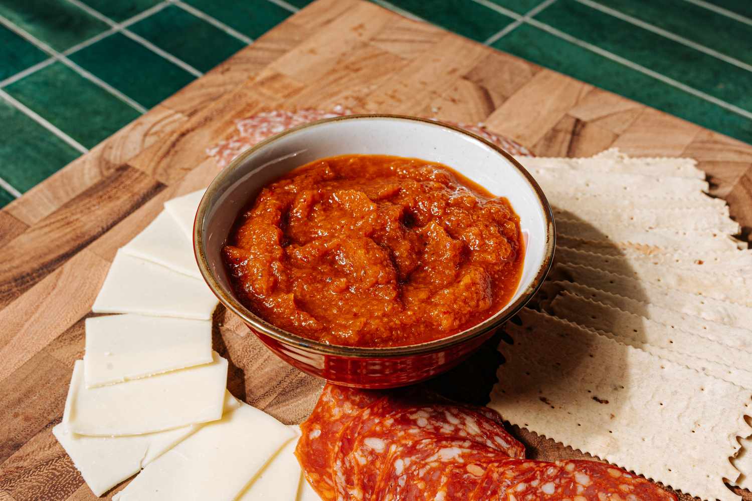 A bowl of red sauce surrounded by sliced cheese crackers and cured meat on a wooden surface