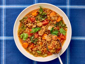 A bowl of sausage and lentil soup with vegetables garnished with parsley on a blue plaid tablecloth